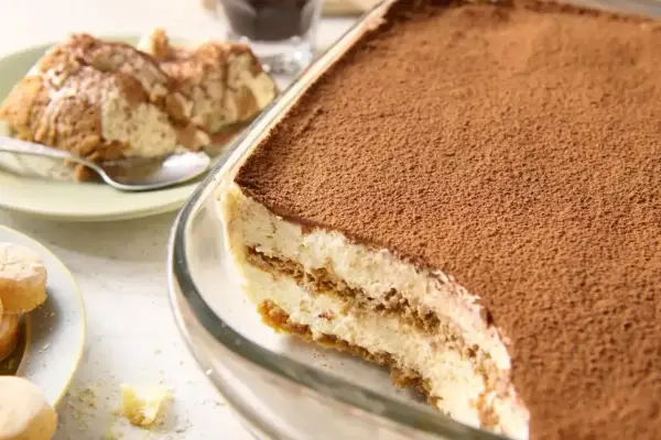 Pyszne tiramisu bez jajek w szklanej formie, posypane kakao. Obok porcja deseru na talerzyku.